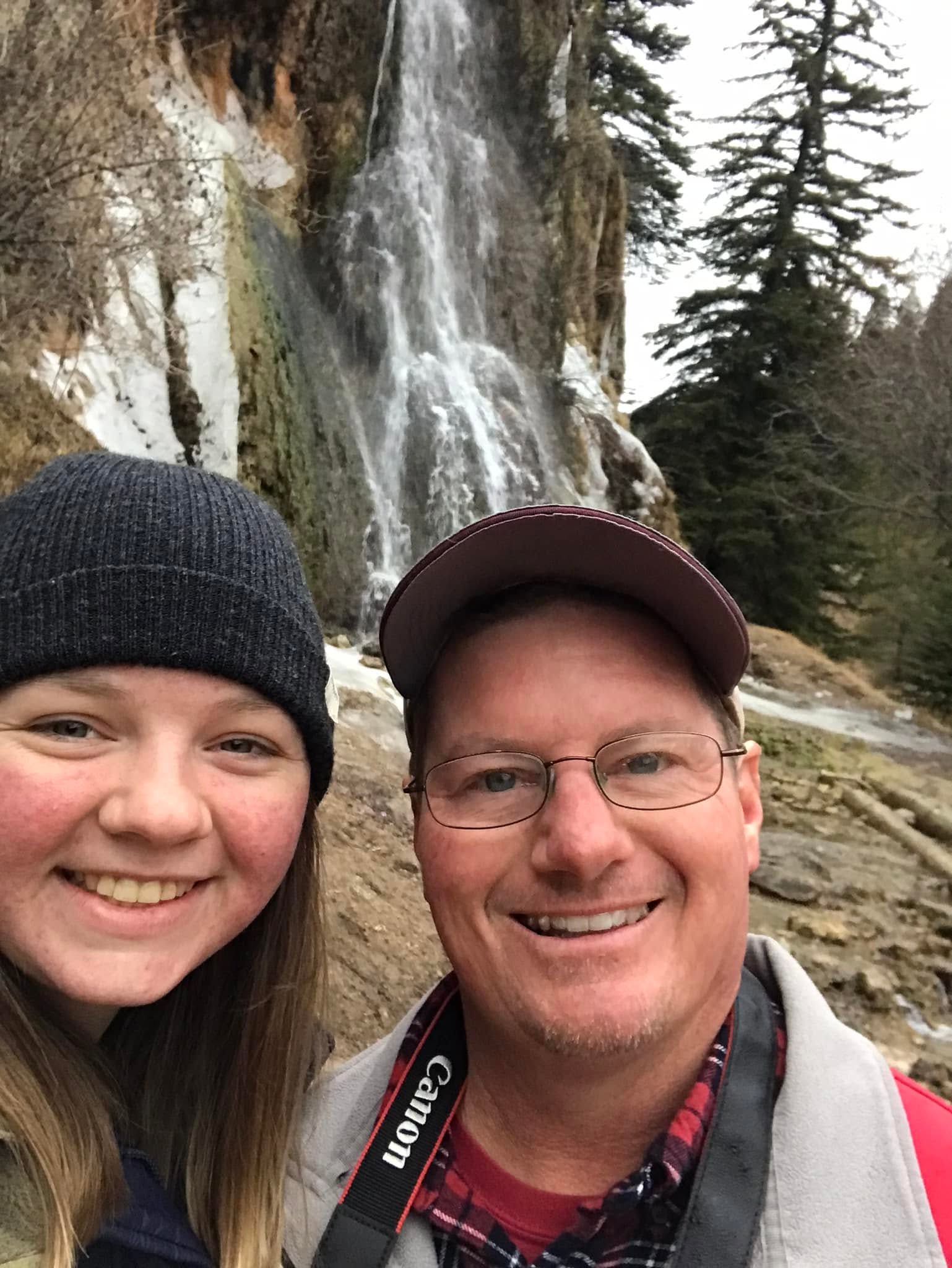 Two people smiling for a photo beside a waterfall.